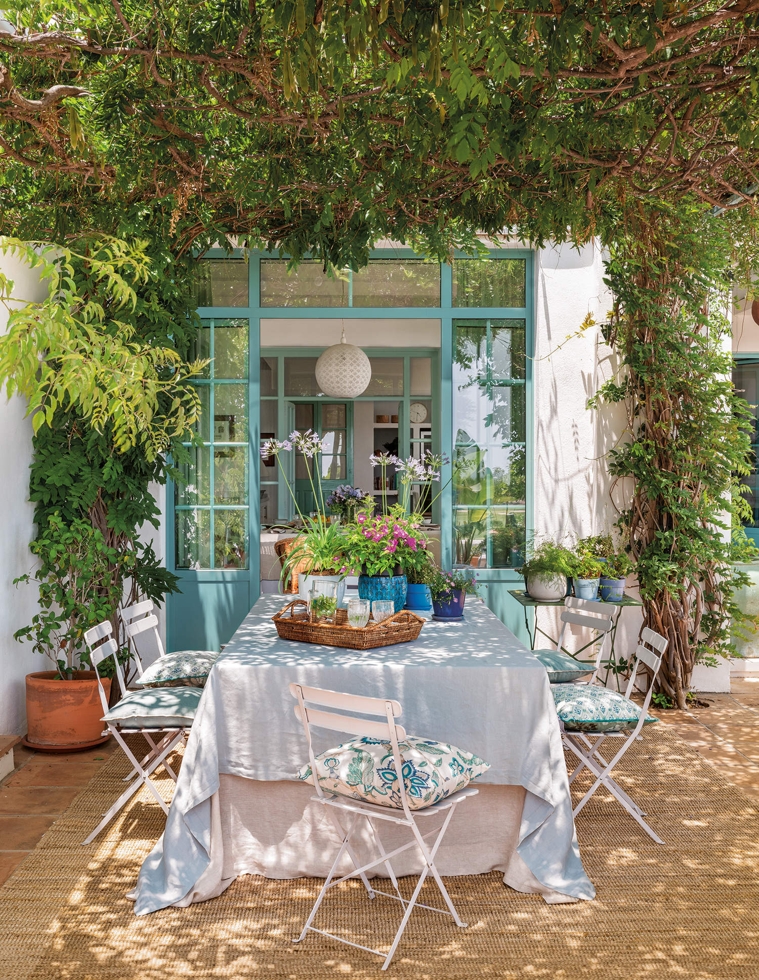Porche fresco con comedor, plantas, mesa y sillas de hierro blanco con cojín azul y alfombra de yute